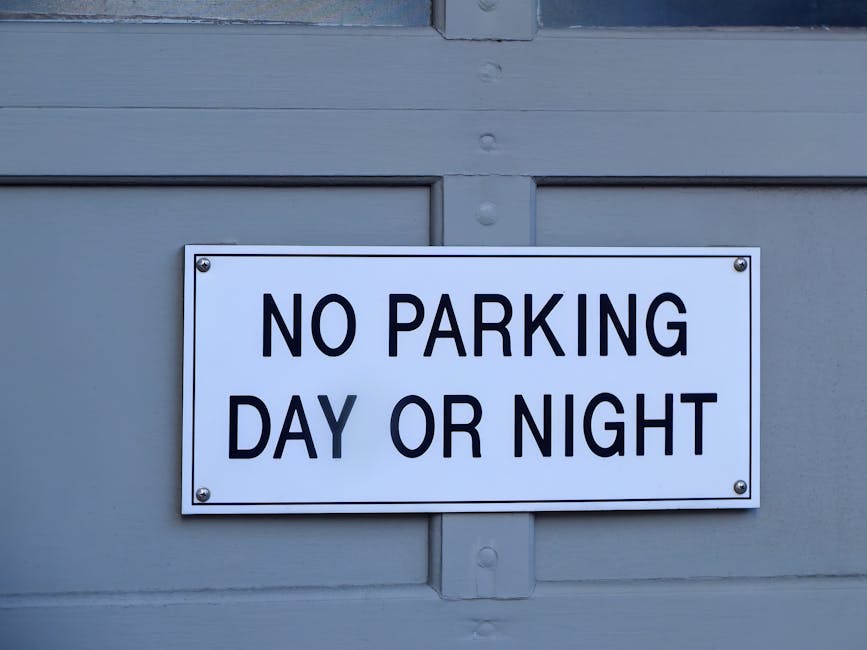 A rectangular white sign with bold black lettering reading 'NO PARKING DAY OR NIGHT' is mounted on a light grey surface, likely a garage door or building exterior. The sign is secured with four silver screws, one at each corner. The background shows horizontal and vertical paneling, indicating a structured, painted surface typical of residential or commercial properties. In the context of home relocation or furniture transport, this sign indicates parking restrictions in the area where the loading or unloading of furniture and boxes by a Man with Van Fulwell crew might be planned. The setting appears to be outdoors, with natural lighting illuminating the sign and surface, emphasizing the importance of adhering to parking rules during moving or packing activities to avoid fines or disruptions.