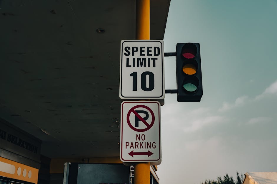 Close-up view of a street traffic signs mounted on a yellow pole at the edge of a pavement, including a speed limit sign indicating a maximum of 10 miles per hour, a vertical traffic light showing red, yellow, and green signals, and a 'No Parking' sign with a red circle and slash over a letter 'P' accompanied by the text 'No Parking.' The background features part of a building with a dark overhang, and the sky above is partly cloudy. This scene depicts urban infrastructure related to parking restrictions and traffic management, relevant to house removals and moving services by Man with Van Fulwell, situated in Fulwell, Richmond.