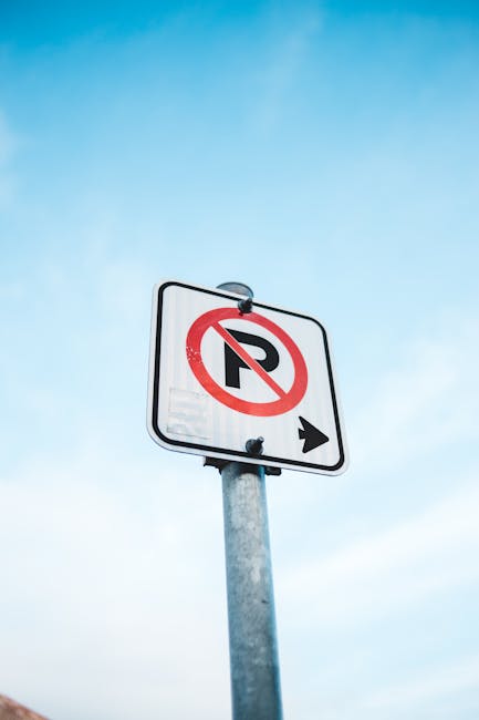 A rectangular white sign with bold black lettering reading 'NO PARKING DAY OR NIGHT' is mounted on a light grey surface, likely a garage door or building exterior. The sign is secured with four silver screws, one at each corner. The background shows horizontal and vertical paneling, indicating a structured, painted surface typical of residential or commercial properties. In the context of home relocation or furniture transport, this sign indicates parking restrictions in the area where the loading or unloading of furniture and boxes by a Man with Van Fulwell crew might be planned. The setting appears to be outdoors, with natural lighting illuminating the sign and surface, emphasizing the importance of adhering to parking rules during moving or packing activities to avoid fines or disruptions.