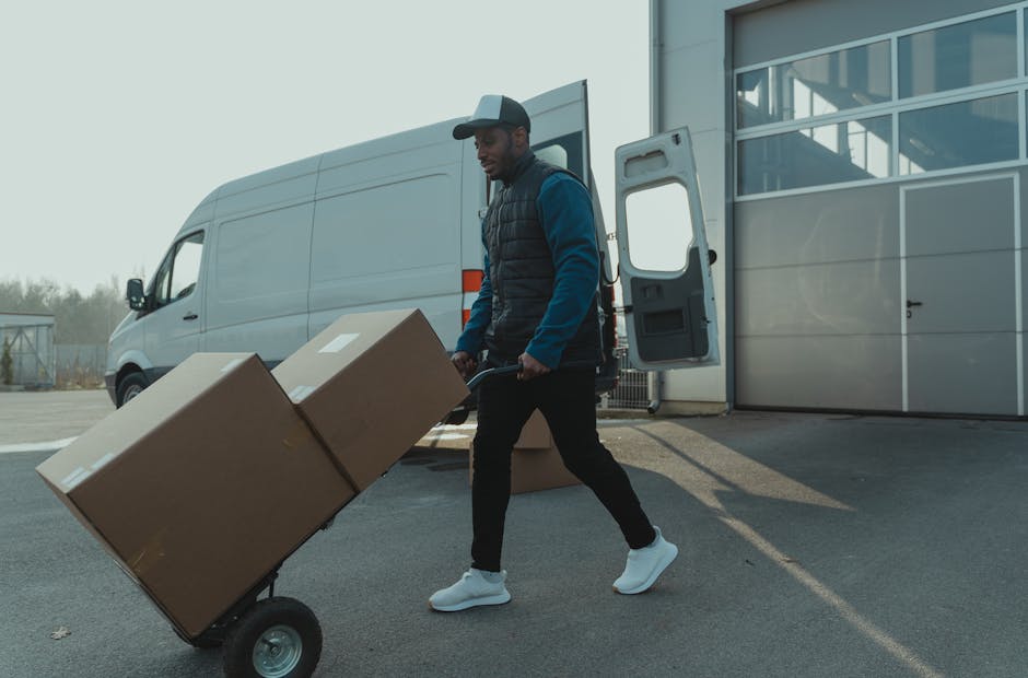 A silver delivery van parked on a residential street during daytime, with a smiling man sitting in the driver's seat and looking out of the open window. The van bears the text 'DELIVERY ALWAYS ON TIME