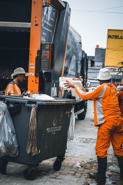 A worker wearing an orange high-visibility uniform and a white helmet is loading a cardboard box into the back of a large black and orange waste collection truck parked on a paved street. Another worker, dressed similarly, stands nearby, assisting with the loading process. The vehicle is positioned next to a building with a yellow sign in the background that reads 'PATHLAB.' The scene takes place in an urban area during daylight, with additional people and signage visible in the background. This image depicts a waste or bulky item collection activity, which is part of the logistics involved in house removals and relocation services as offered by Man with Van Fulwell. The operation involves careful handling of packaging materials such as cardboard, and the use of equipment like the truck for efficient furniture transport and packing and moving during a house relocation.