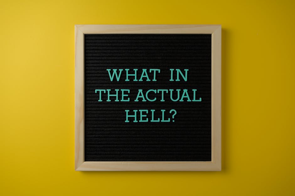 A black felt letter board with a light wooden frame is mounted on a bright yellow wall. The board displays the message 'WHAT IN THE ACTUAL HELL?' in uppercase teal letters. The image suggests an element of exaggeration or frustration, which may relate to the complexities of home relocation or moving logistics, as seen in the context of house removals and packing by Man with Van Fulwell. The lighting is even, highlighting the contrast between the dark board and the vibrant background, creating a visually clear and neutral depiction relevant to the theme of moving or handling confusing removal quotes.