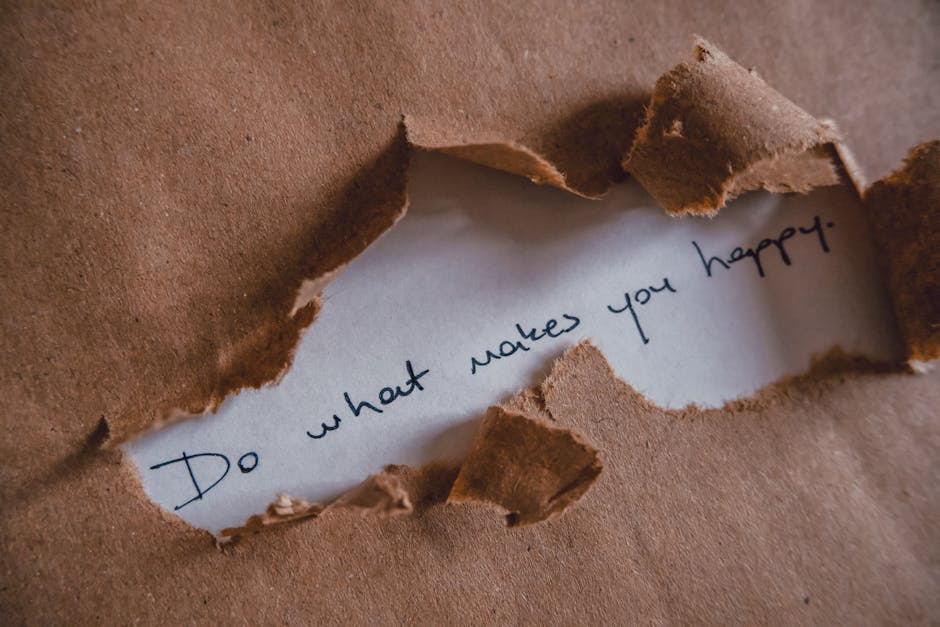 A close-up photograph of a torn piece of brown kraft paper with rough, jagged edges revealing a white inner surface where the handwritten message 'Do what makes you happy' is visible. The torn paper is placed on a flat surface, likely part of a packing or moving process, suggesting careful handling or packing materials used during home relocation. The lighting is natural, illuminating the texture of the paper and the handwritten text in dark ink. The background is out of focus, indicating an indoor setting associated with moving or packing activities, and subtly supporting the context of furniture transport and packing during house removals, as provided by Man with Van Fulwell.