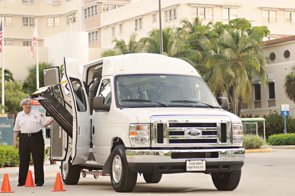 A white Ford Transit van is parked outside a residential building on a paved area surrounded by greenery, including palm trees and bushes. The van's side door is open, revealing the interior with furniture and boxes arranged for loading or unloading. A man wearing a white shirt and dark trousers stands beside the van, holding the open door, indicating a home relocation or furniture transport process. There are orange traffic cones placed near the van, suggesting an organized loading zone, and the overall scene is well-lit with natural daylight. The background features apartment buildings and a sign indicating accessible parking, all contributing to a setting focused on moving services, as provided by Man with Van Fulwell.