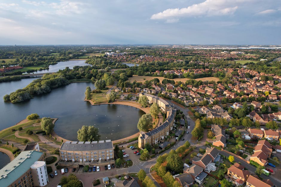 An aerial view of Fulwell Park Estate showing a curved residential area with terraced and detached houses, some with driveways and garages, situated around a large, irregularly shaped lake. The lake features a small island with trees and a paved walkway around its perimeter. Adjacent to the residential units, there is a parking lot with numerous parked cars, and a pathway lined with trees connects different parts of the estate. In the background, the landscape extends to open fields and additional housing, under a partly cloudy sky. The scene captures the outdoor environment commonly involved in home relocation and furniture transport, as viewed during the loading process carried out by Man with Van Fulwell, demonstrating the spatial arrangement of residential properties, green spaces, and access points necessary for effective removals services.