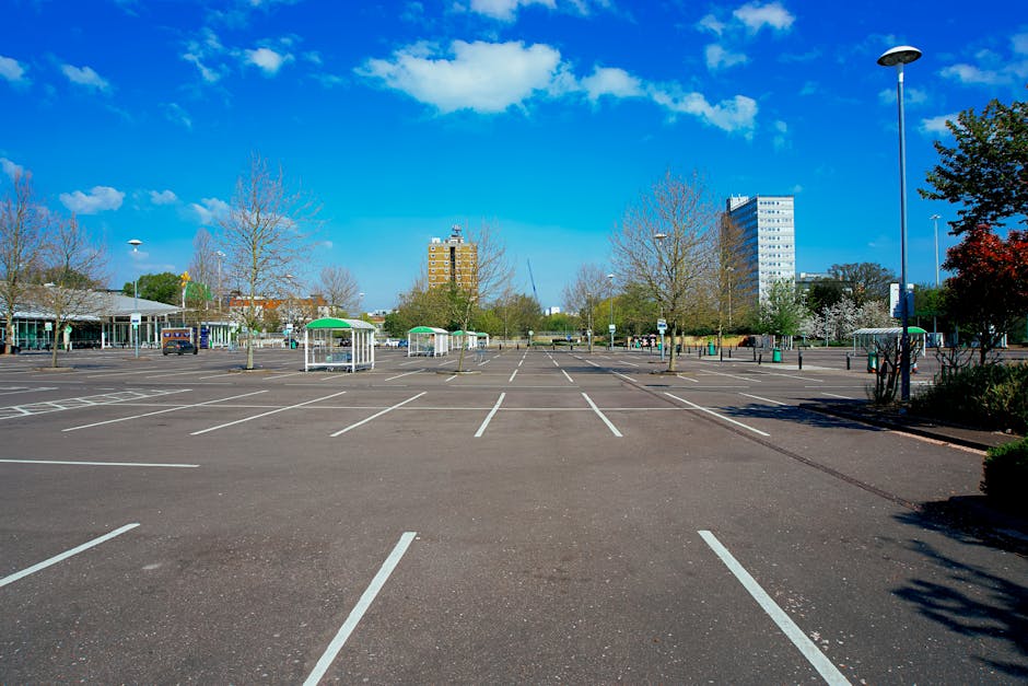 A vacant outdoor parking lot with multiple empty parking spaces marked by white lines on dark asphalt, situated in a residential or commercial area during daylight with bright blue skies and scattered clouds overhead. Several leafless and partially blossoming trees are visible along the edges of the lot, along with a few small, sheltered street stalls or kiosks with green roofs positioned toward the center. A few parked cars are seen in the distance, and modern multi-storey office or apartment buildings rise behind the trees, indicating urban surroundings. Street lighting poles and some signage are also present within the parking area. This setting depicts a typical location where a home relocation or furniture transport company like Man with Van Fulwell might coordinate parking arrangements for efficient loading or unloading during removals, showing the spacious and organized environment suitable for moving logistics, including the use of trolleys, straps, or vans for interior or exterior furniture transport.