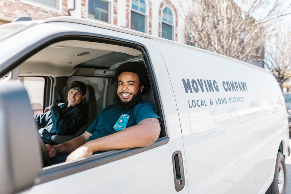 Inside a white moving van registered to Man with Van Fulwell, two movers are seated in the vehicle during a home relocation. The driver, a man with dark curly hair, a beard, and wearing a dark blue T-shirt, is smiling at the camera, with his arm resting on the door frame, situated in the front seat. Sitting behind him is a woman with dark hair tied back, wearing a black and grey jacket, also looking towards the camera with a cheerful expression. The van’s interior reveals cardboard boxes and packing materials, such as plastic wrap and blankets, indicating an active packing and moving process. Outside the open passenger door, the driveway or street is visible, with a building and some leafless trees in the background, suggesting a residential area during mild weather. The vehicle displays the company’s branding and tagline, emphasizing their local and long-distance relocation services, supporting the context of furniture transport and packing during a house move in Fulwell.