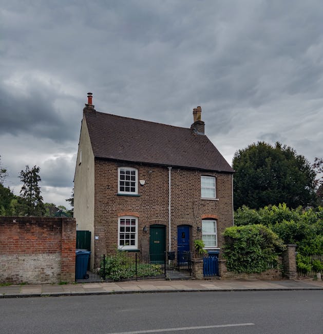 A two-storey semi-detached house with a brick facade and a pitched roof, situated on a residential street under a cloudy sky. The house has four white-framed sash windows—two on the upper floor and two on the ground floor—along with a green front door and a blue second door. In front of the house, there is a paved footpath and a small garden area with some green bushes and plants. Adjacent to the house, a low brick wall separates the property from the sidewalk, which features two blue wheelie bins. The street is lined with other greenery and trees, and the overall scene depicts a typical suburban home exterior, as might be encountered during a home relocation or furniture transport process coordinated by a professional removals service such as Man with Van Fulwell.