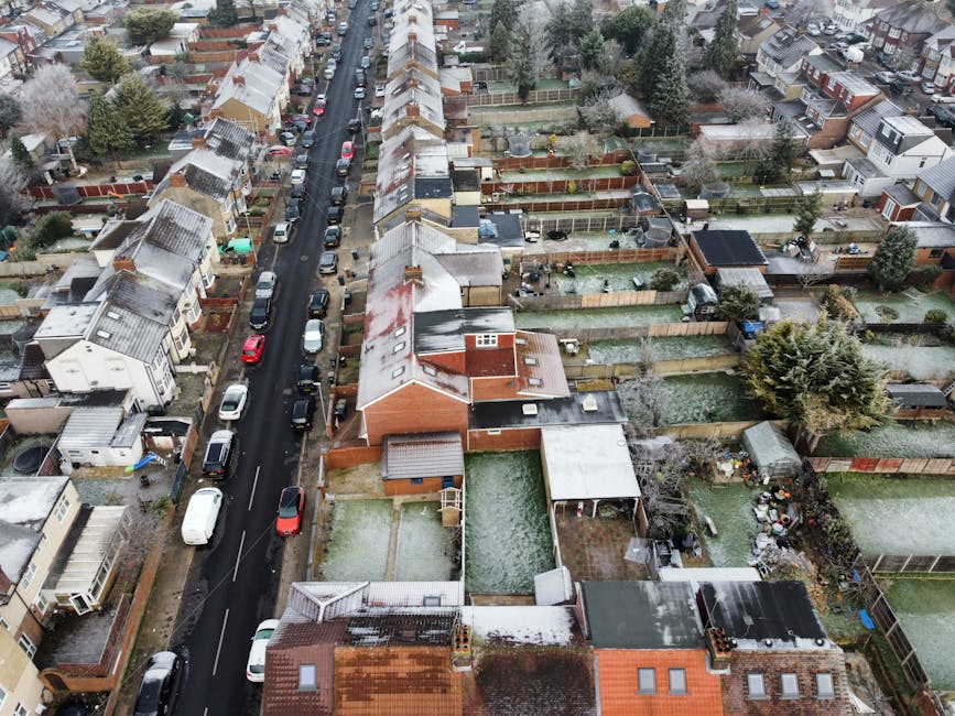 An aerial view of a residential street covered with a light dusting of snow, showing rows of terraced houses with pitched roofs, some of which have snow accumulation. The street is lined with parked cars on both sides, including sedans, hatchbacks, and vans, with varied colours such as white, red, and black. Each property has a backyard enclosed by wooden fences, many featuring sheds, garden furniture, and potted plants. Some gardens have visible lawn areas, patios, or paved sections, with a few containing garden tools or toys. The houses display diverse exterior materials, including brick, weatherboard, and painted surfaces. The lighting appears diffused under an overcast sky, typical of winter weather. This scene illustrates the typical residential environment that Man with Van Fulwell might work within for house removals or furniture transport, especially considering the space for loading vehicles in the street for home relocation or packing and moving logistics connected to Richmond Council rules on house moves and skip permits.