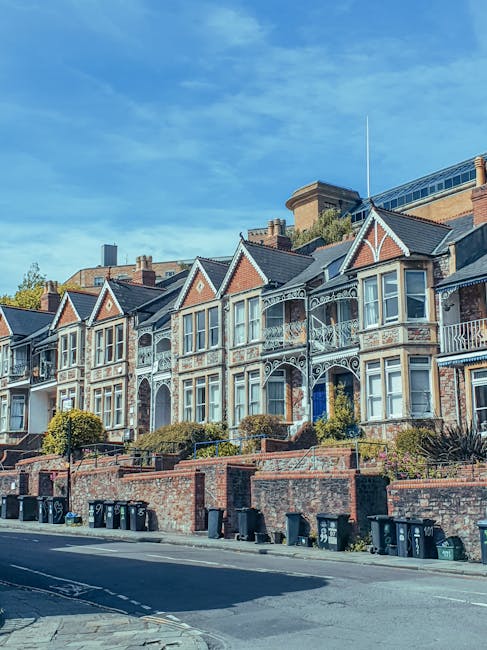 A two-storey semi-detached house with a brick facade and a pitched roof, situated on a residential street under a cloudy sky. The house has four white-framed sash windows—two on the upper floor and two on the ground floor—along with a green front door and a blue second door. In front of the house, there is a paved footpath and a small garden area with some green bushes and plants. Adjacent to the house, a low brick wall separates the property from the sidewalk, which features two blue wheelie bins. The street is lined with other greenery and trees, and the overall scene depicts a typical suburban home exterior, as might be encountered during a home relocation or furniture transport process coordinated by a professional removals service such as Man with Van Fulwell.