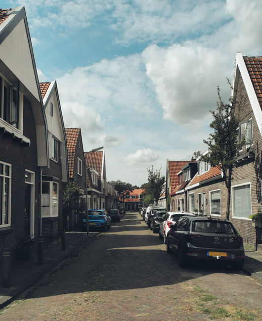 A narrow residential street with terraced houses on either side featuring brick and painted facades, some with small front gardens and potted plants. Several cars, including a dark hatchback and a lig