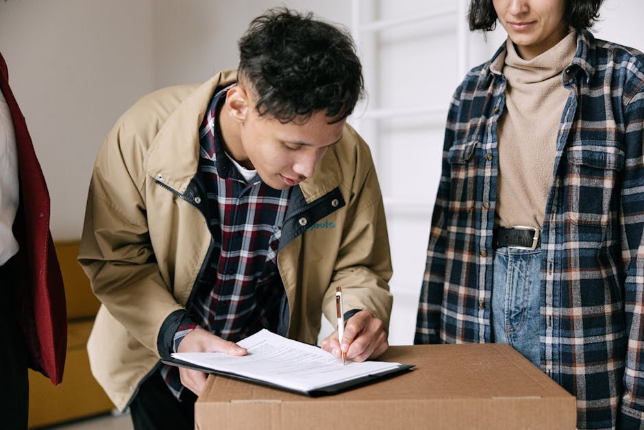 A young man with dark, curly hair wearing a beige outdoor jacket over a checkered shirt is seen signing a delivery or inventory document on a clipboard, placed on top of a cardboard box. He is inside a home, next to another person wearing a plaid jacket and a beige turtleneck, who stands nearby observing. In the background, there is a staircase with white spindles, indicating an indoor setting. The scene captures a moment during home relocation or furniture transport, as the man with Van Fulwell prepares to load or unload items, with the cardboard box ready for moving logistics. The lighting is natural, highlighting the details of the packing materials and the expressions of the individuals involved in the packing and moving process.