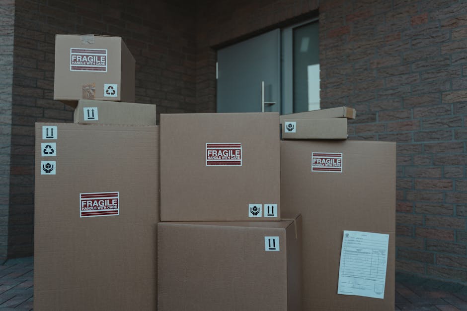 Several cardboard boxes of varying sizes filled with household items are stacked outside a house near a grey door. The boxes are sealed with packing tape and feature labels indicating fragility and handling instructions, such as 'Fragile handle with care,' along with symbols for glass, recycling, and proper orientation. Some boxes are placed on the pavement, while others are stacked directly against the exterior brick wall of the property. The scene appears to be part of a home relocation process, with boxes prepared for safe furniture transport and packing and moving activities. The background includes a portion of the brick house exterior and a door with a glass panel. The presence of these packed boxes suggests the early stages of a house removal, possibly handled by [COMPANY_NAME], which specializes in removals and moving services, as implied by the context of the associated webpage focused on urgent Fulwell same-day moves.