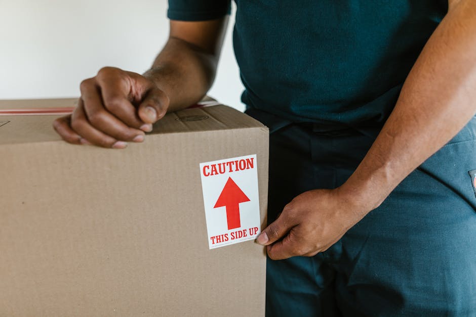 Close-up of a person wearing a dark shirt and blue trousers securing a large cardboard box with a red and white caution label that reads 'CAUTION THIS SIDE UP.' The individual is holding the box firmly, with one hand on the side and the other on top, indicating preparation for a home relocation or furniture transport. The cardboard box appears sturdy, with visible packing tape, and is positioned on a flat surface, likely indoors, close to a doorway or in an entryway area. The background is neutral, emphasizing the focus on the handling process, which is part of packing and loading during a professional moving service provided by Man with Van Fulwell. The scene illustrates careful handling of goods during the loading process for a prompt, same-day move.
